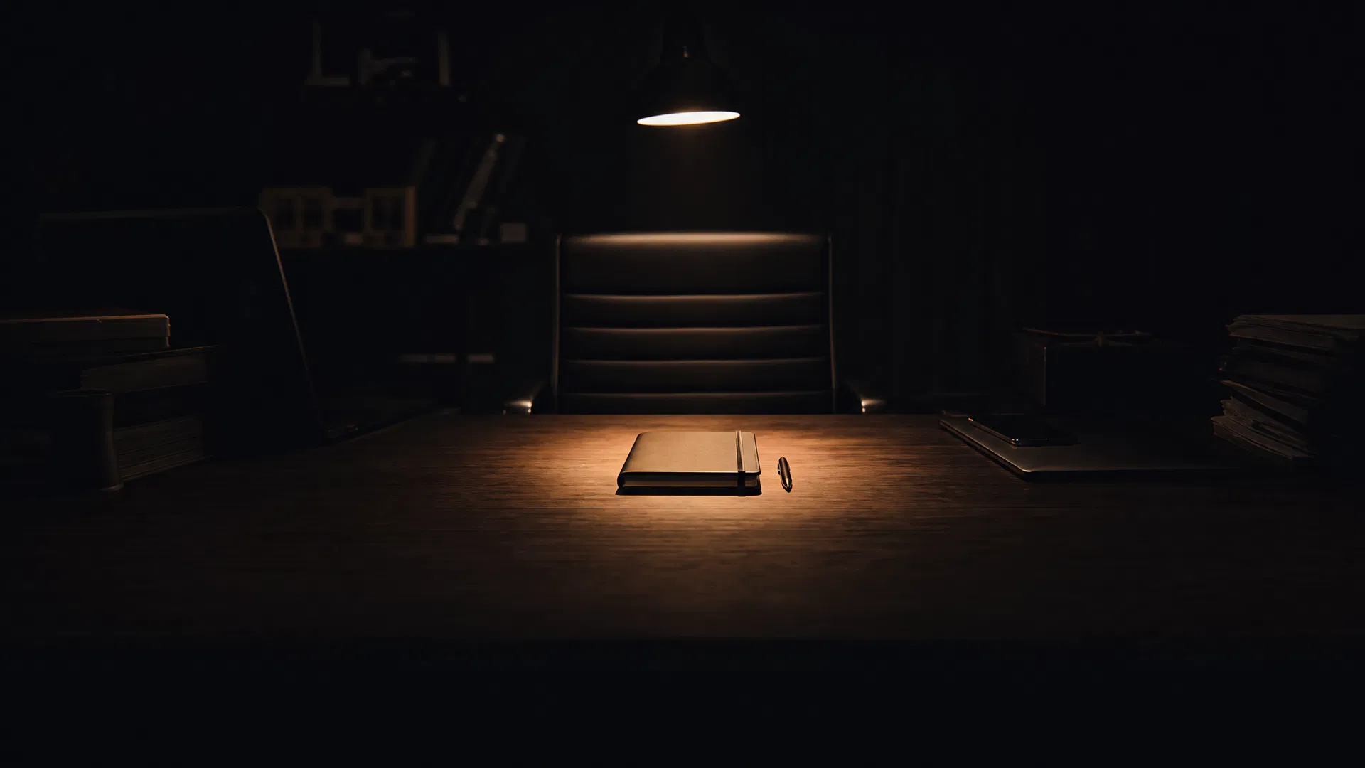 Cinematic dark composition of a minimalist executive desk with a single amber spotlight illuminating one clean notebook and pen at the center, while stacks of papers and cluttered items fade into deep shadow — symbolising strategic focus and the power of subtraction in business scaling